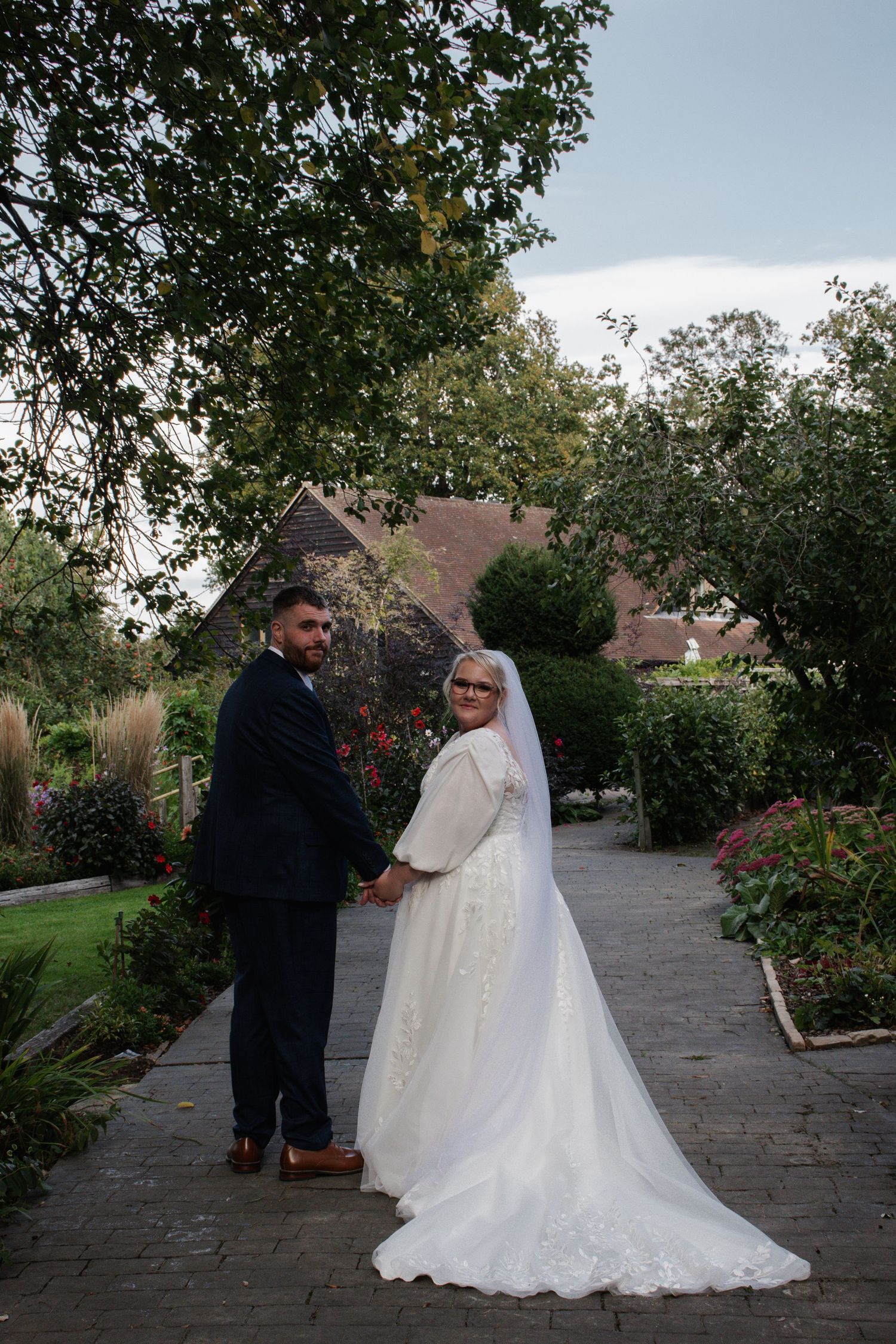 Bride and groom holding hands on a garden pathway, bride in lace gown with long veil and fur wrap.