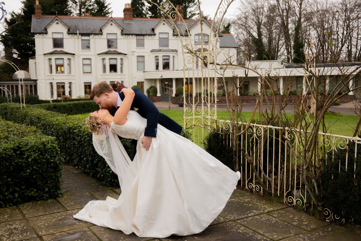 Groom dipping bride in a sweeping white gown outside an elegant country house venue.