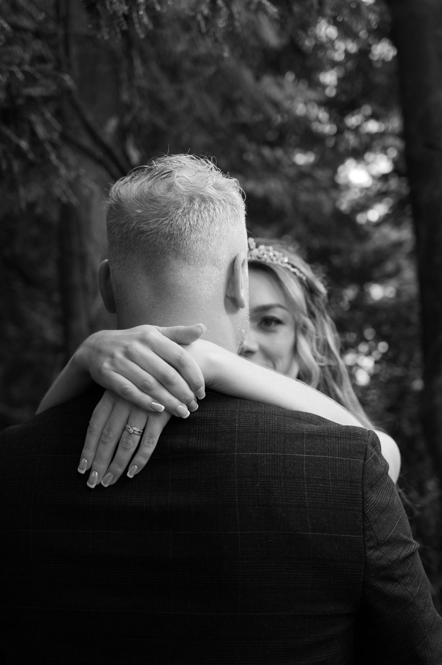 Black and white close-up of bride embracing groom from behind, showing wedding ring on her hand.