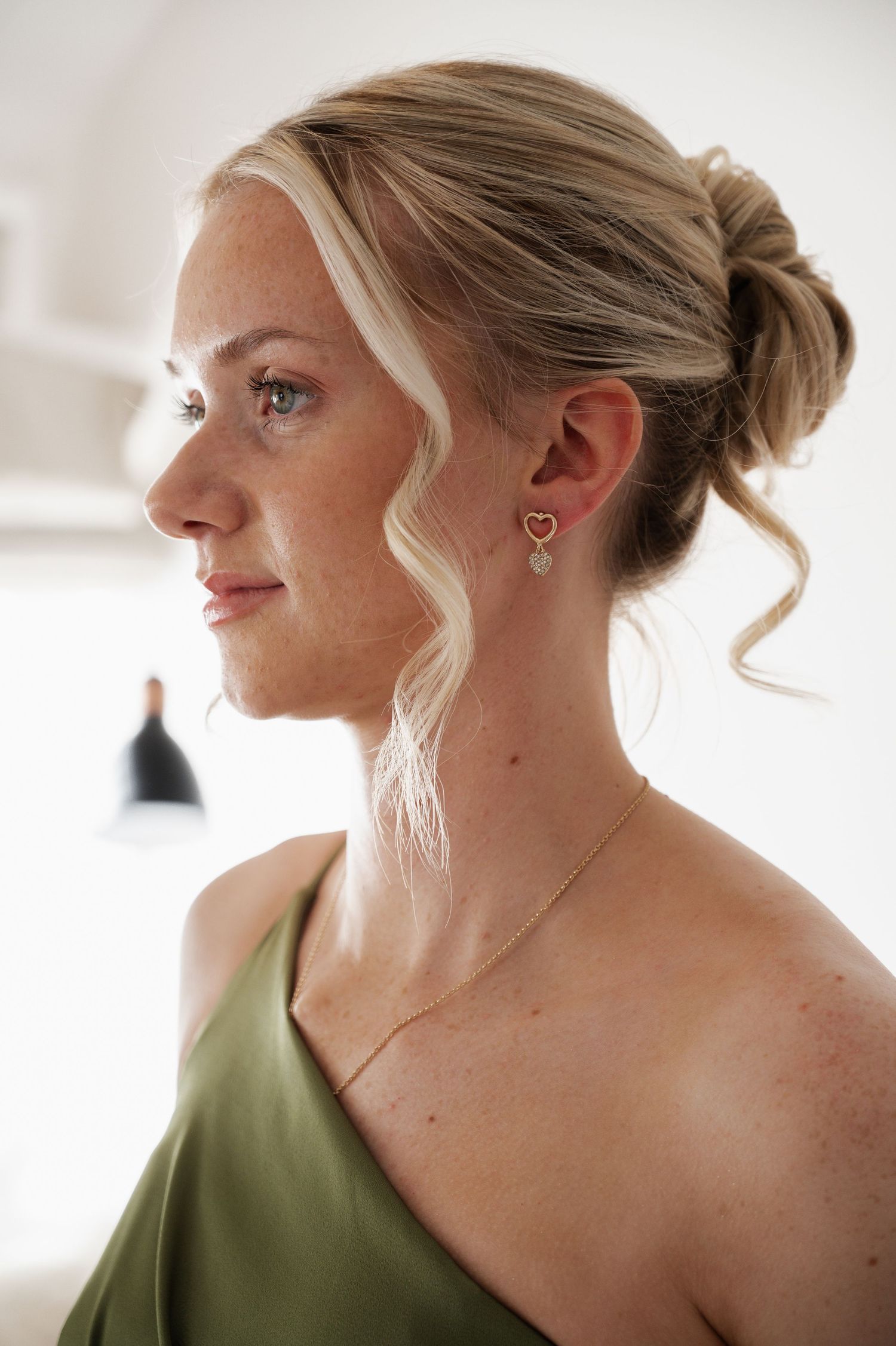 Side profile of an elegant bridesmaid with blonde updo wearing heart earrings and a sage green satin dress.
