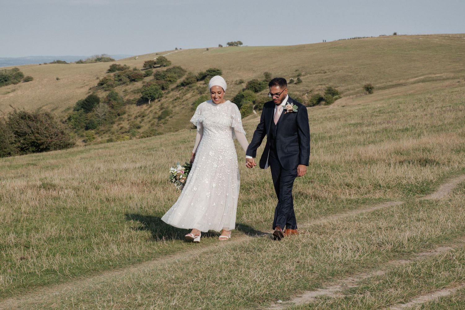 Muslim bride in white hijab and lace gown walks hand-in-hand with groom across a scenic green hillside landscape.