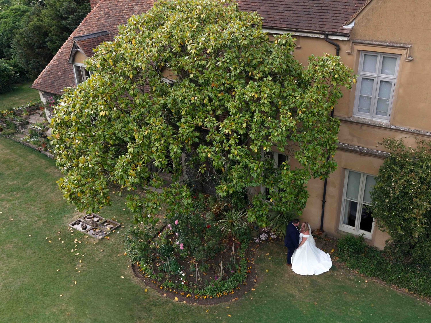 Aerial view of bride and groom embracing beneath a large tree beside a historic stone manor. Nursted Court