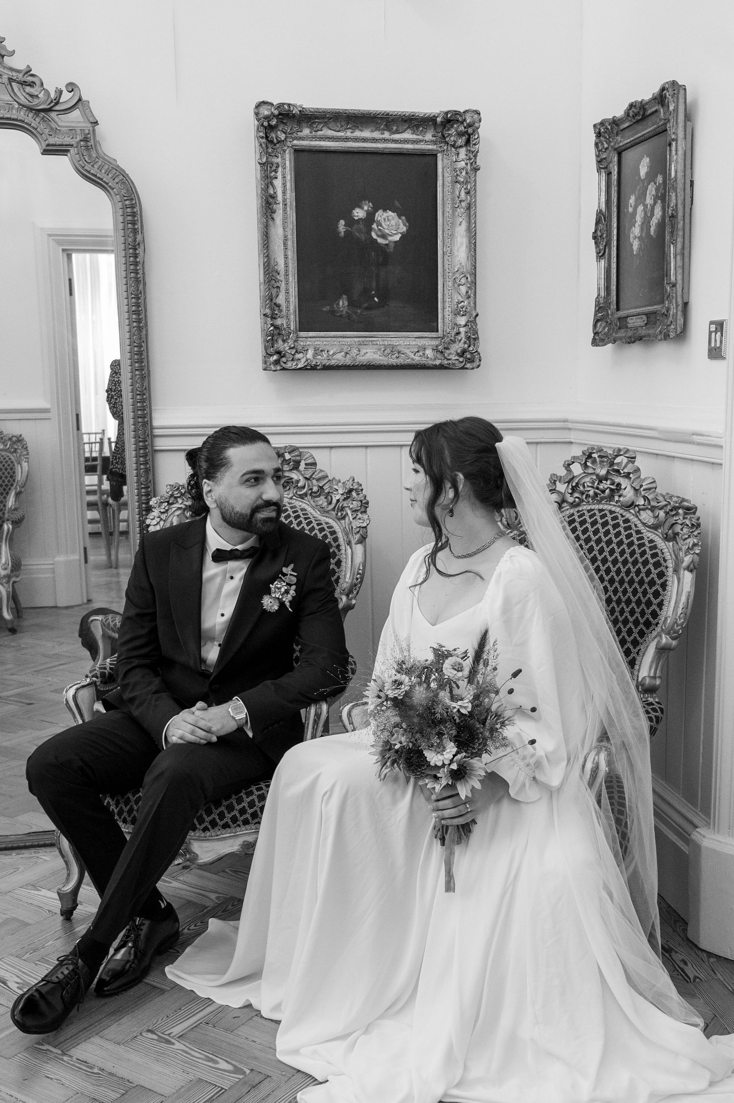 Black and white photo of bride and groom seated on ornate chairs in an elegant room with gilded framed artwork.