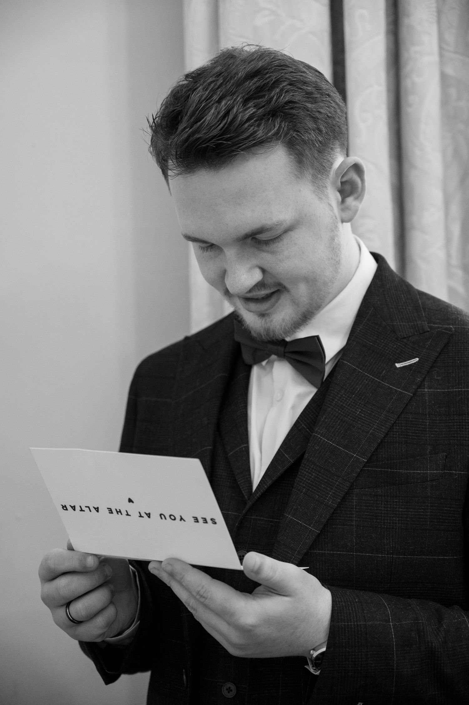 Groom in a black tuxedo reading a see you at the altar card before the ceremony.
