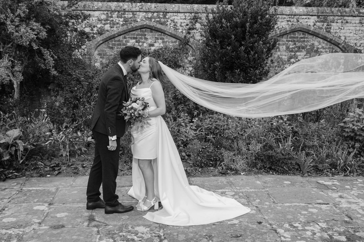 Black and white photo of bride and groom kissing as the wedding veil dramatically flows in the wind.