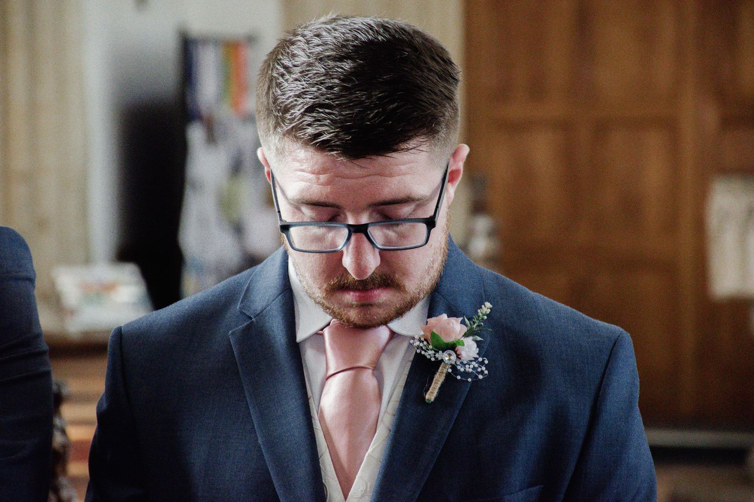 Close-up of a groom in navy suit with pink tie and glasses, looking down with a floral boutonniere at his lapel.