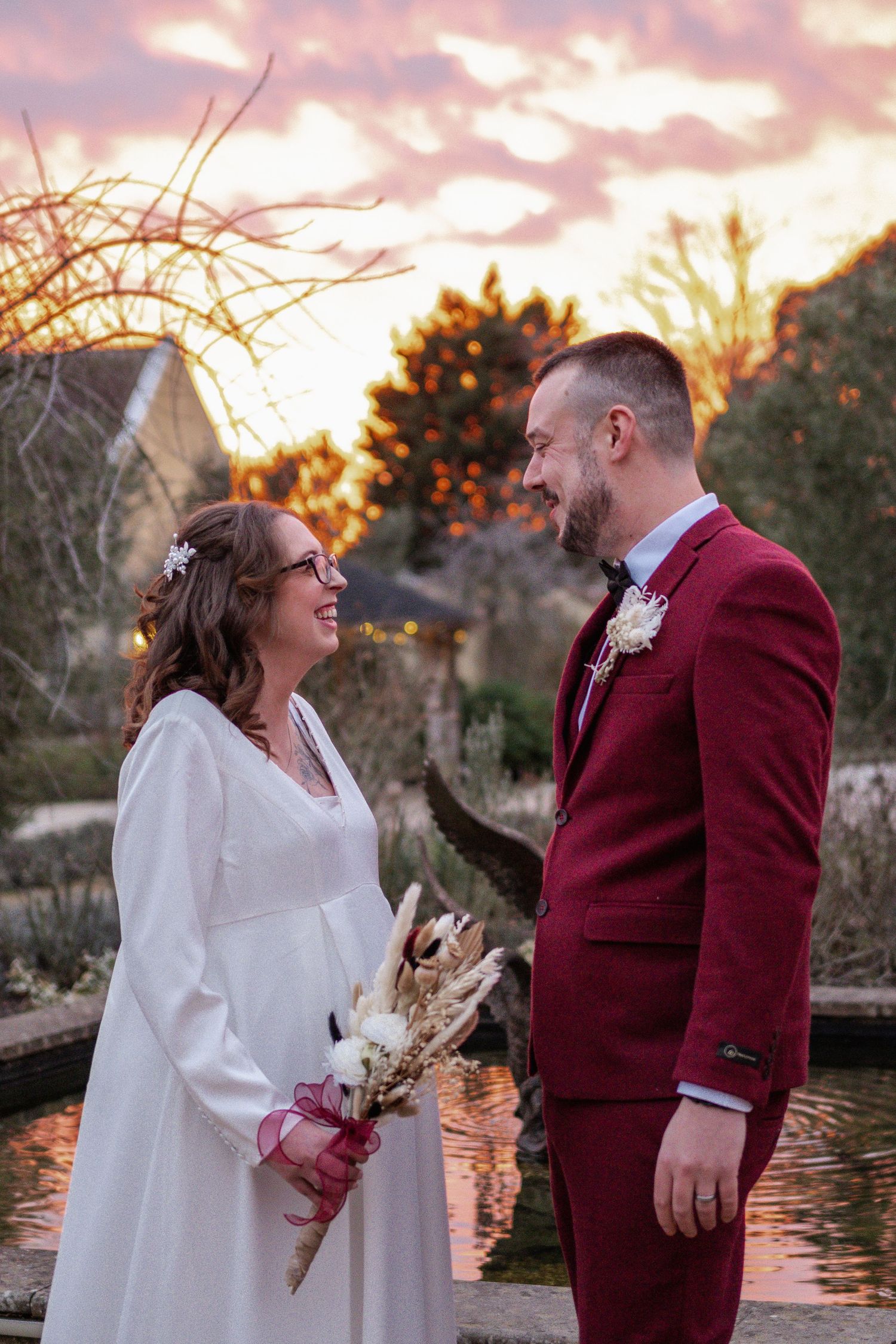 Bride and groom share a romantic sunset moment by a garden pond, she in white gown holding dried bouquet, he in burgundy suit.