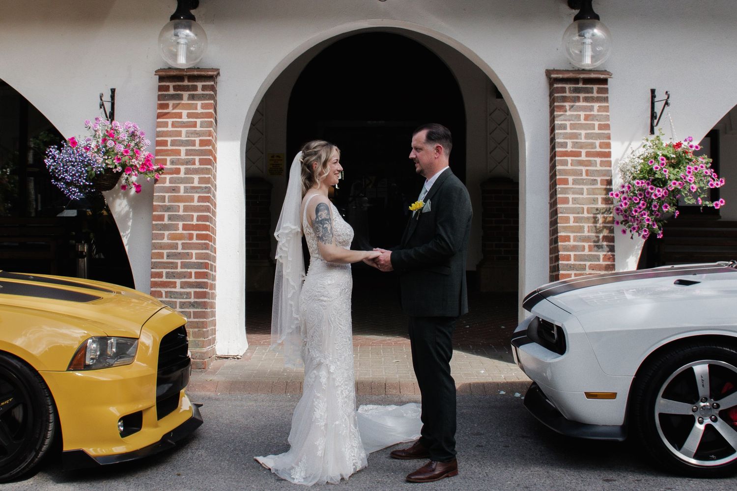 A bride and groom hold hands between two classic muscle cars outside a brick arch venue entrance.