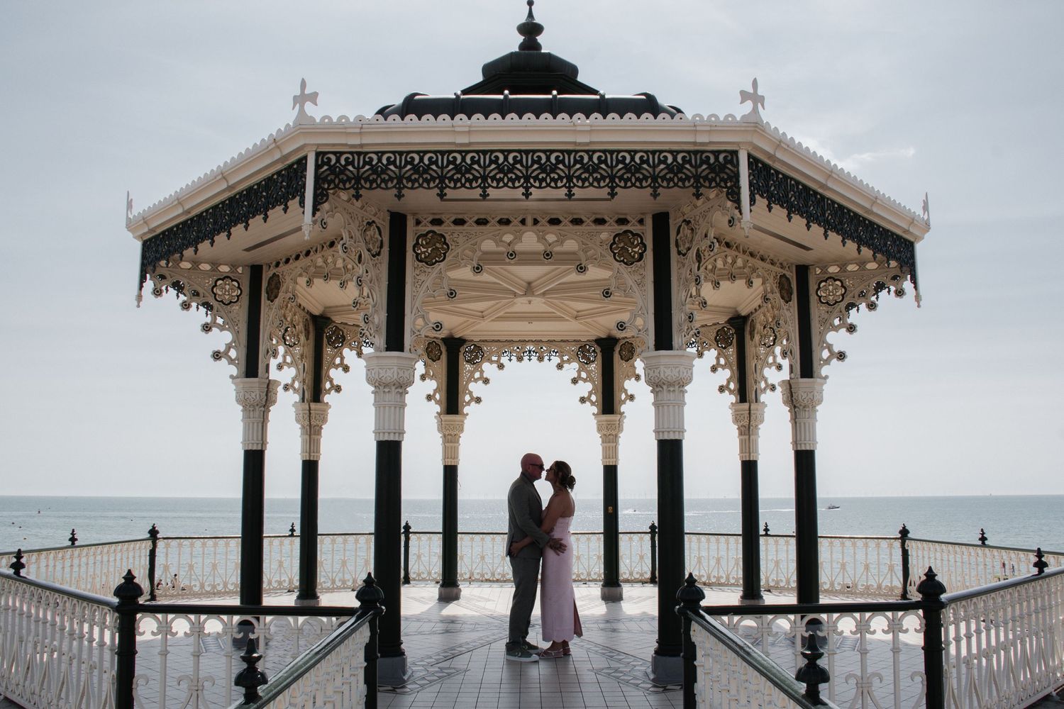 Couple shares romantic embrace under ornate Victorian seaside bandstand with ocean backdrop.