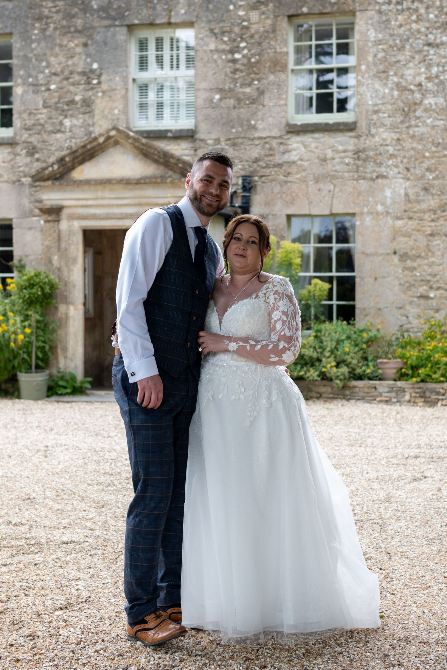 Bride and groom posing together on gravel drive before elegant stone country manor.