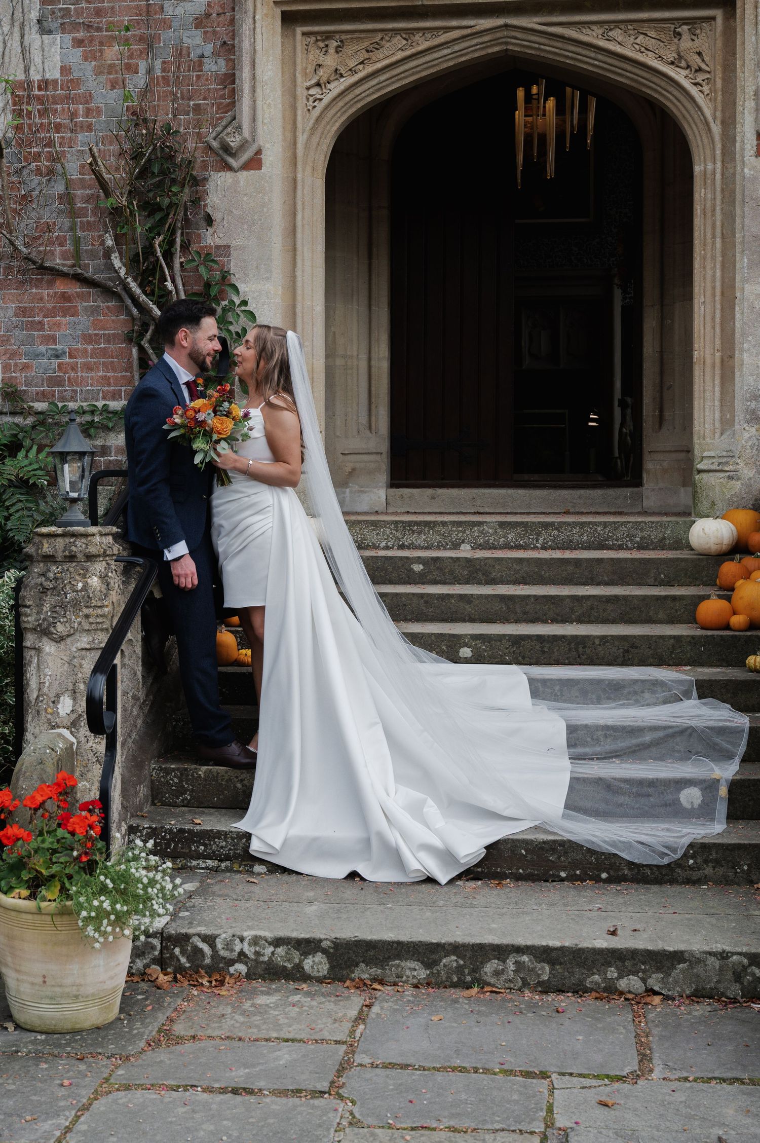 Bride and groom kissing on stone steps of a grand Gothic venue with autumn pumpkins and flowing wedding veil.