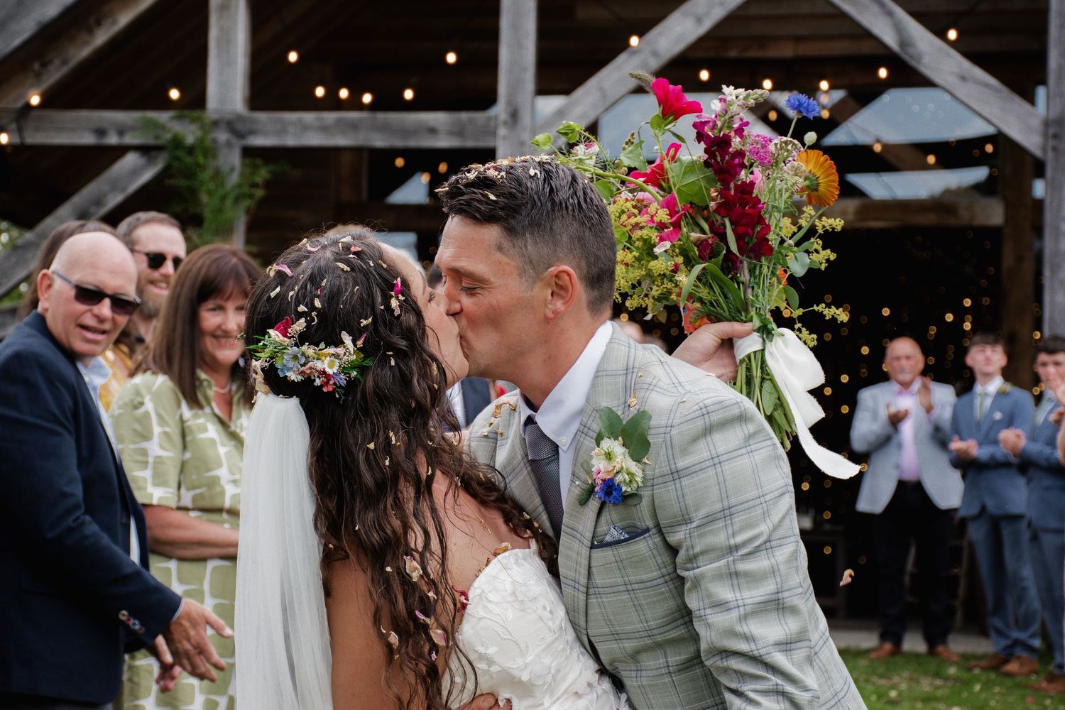 Newlyweds share first kiss at rustic barn wedding while guests celebrate with colorful confetti and floral bouquet.