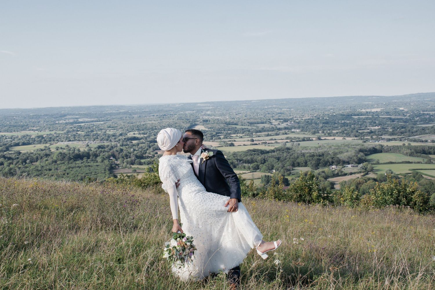 Groom dips bride in white hijab and lace wedding dress on a hilltop with stunning panoramic countryside views below.