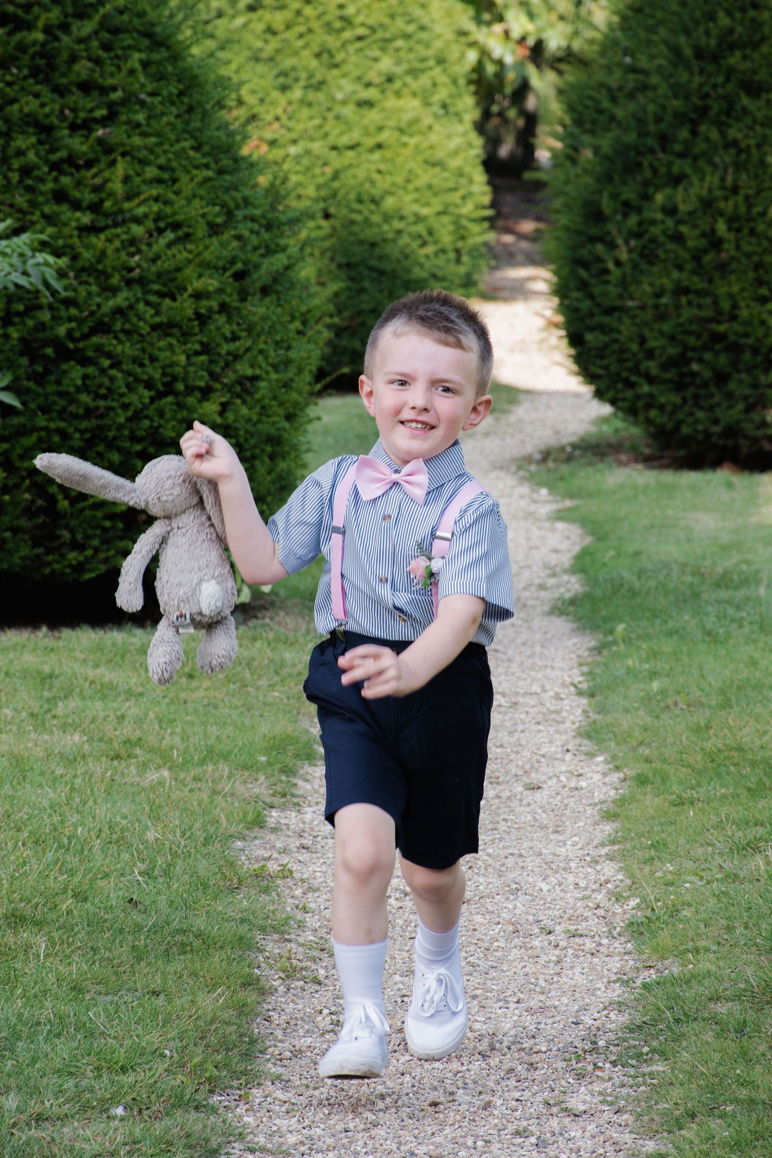 Happy young boy in gray shirt, pink bow tie and suspenders runs along garden path holding a stuffed bunny toy.