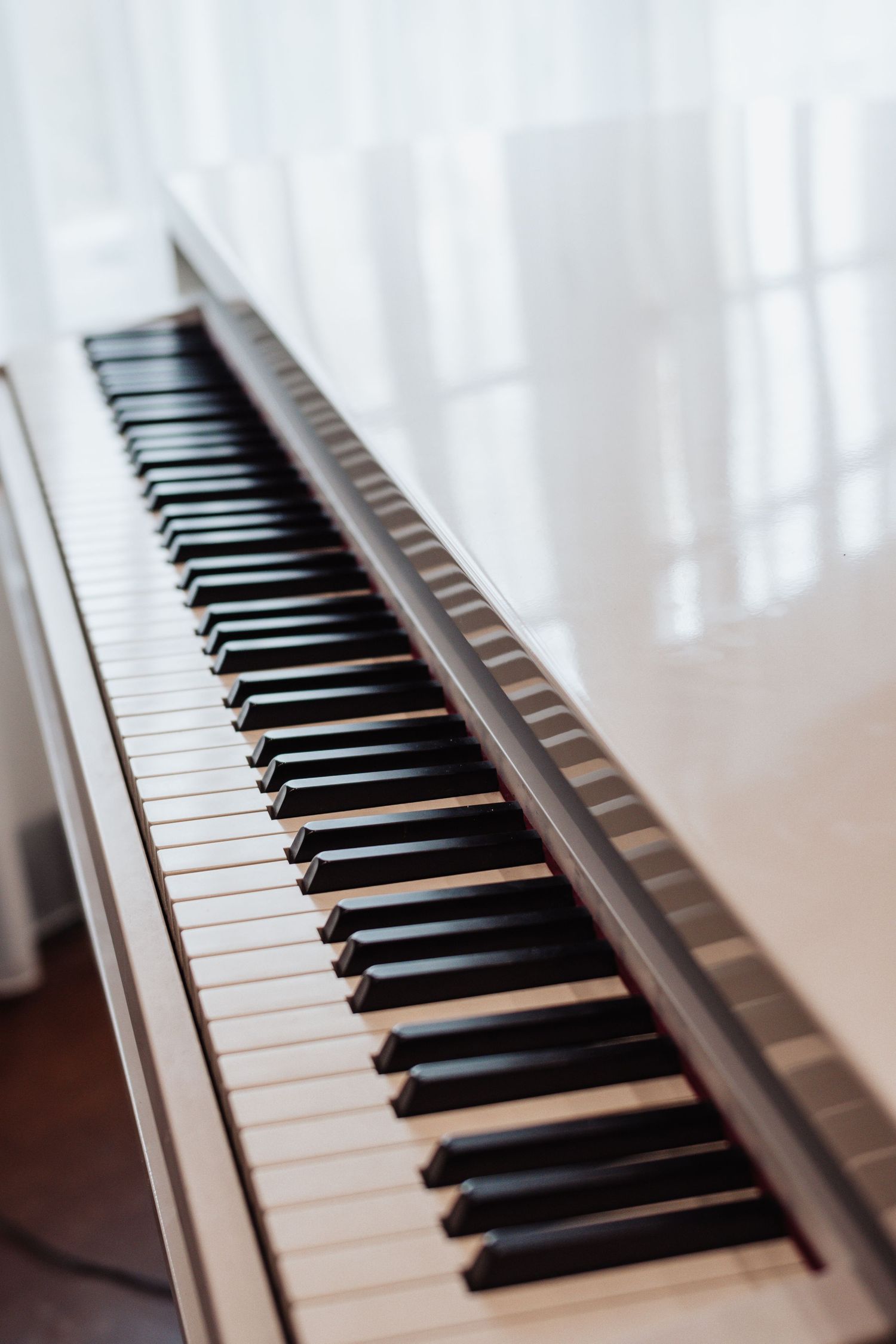 Close-up of elegant white piano keys on a glossy white piano near a bright window with sheer curtains.