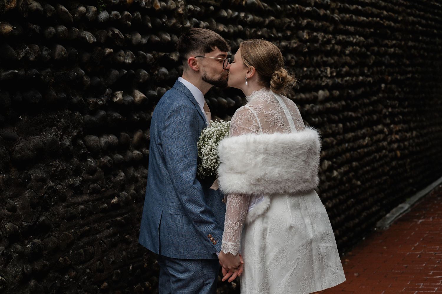 A couple shares a romantic kiss on their wedding day, bride in white fur stole, groom in blue suit.