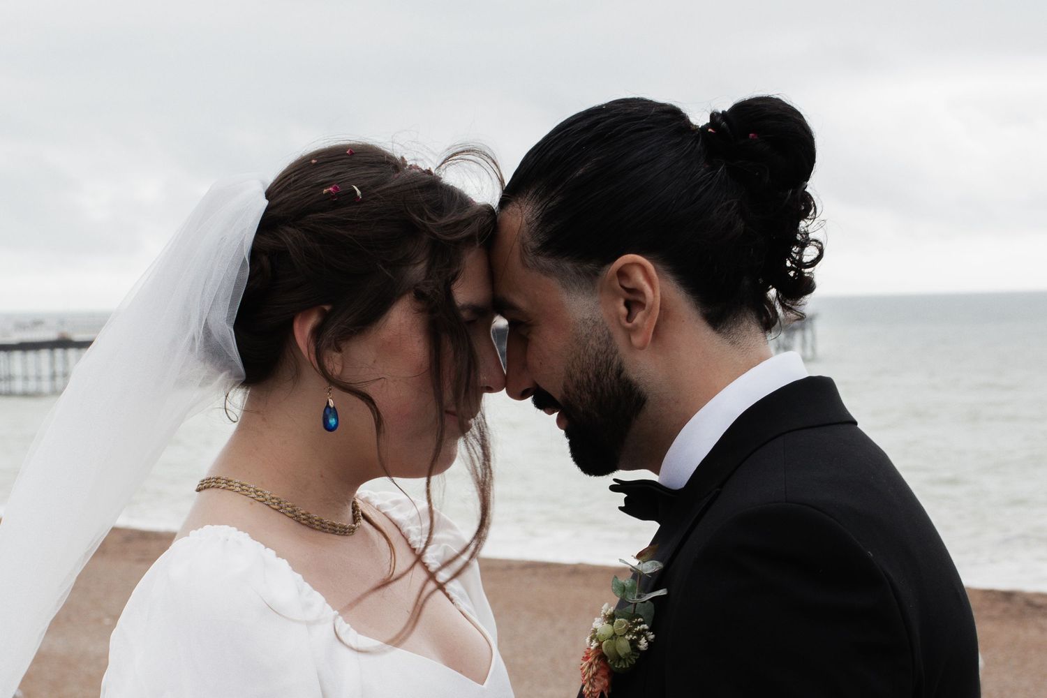 Bride and groom share an intimate forehead touch on a cloudy beach, with a distant pier in the background.