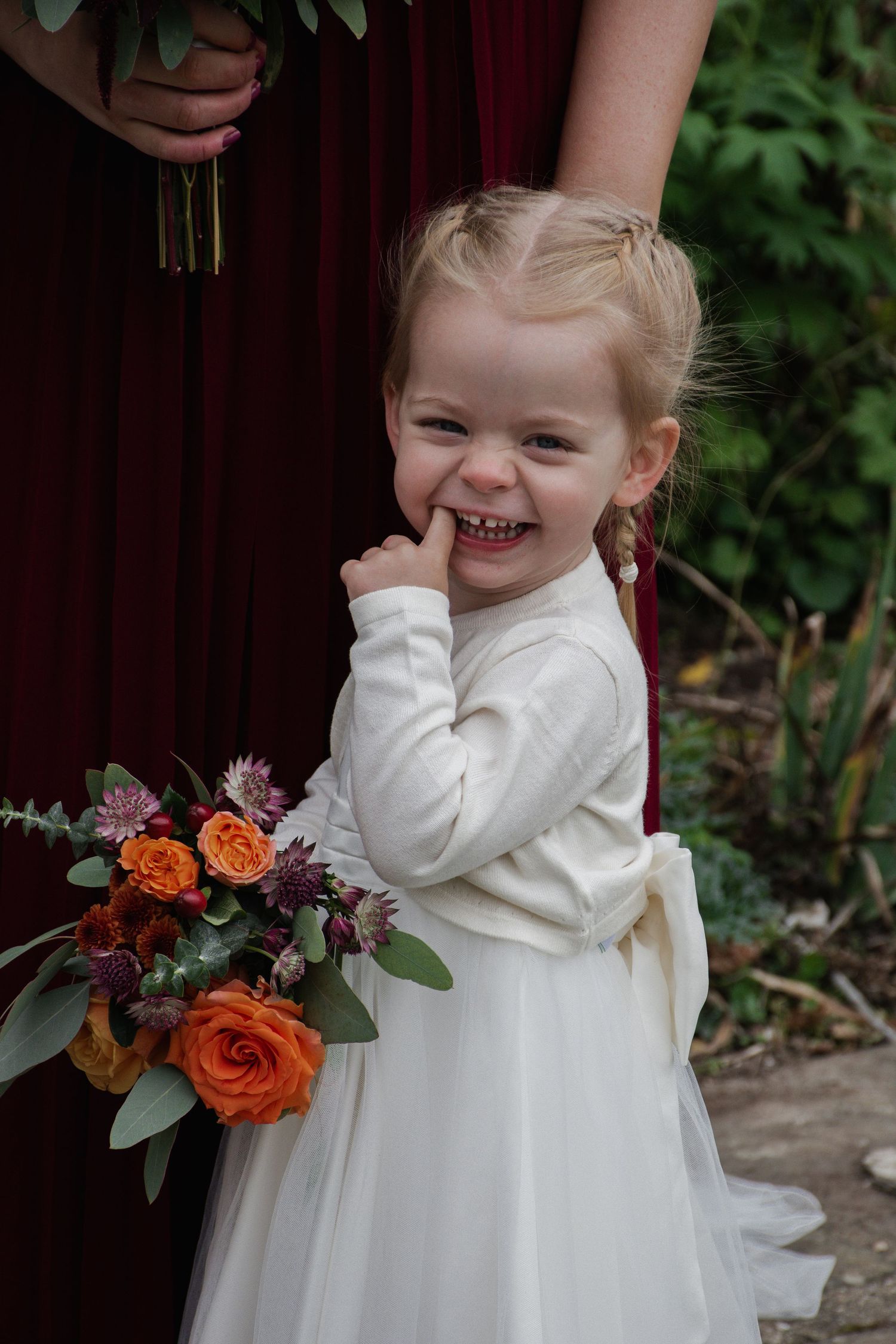 Adorable smiling flower girl in white dress holding a bouquet of orange roses at an autumn wedding.