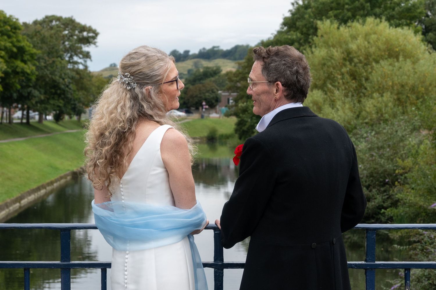 Wedding couple stand hand in hand on a blue bridge overlooking a peaceful river, bride wearing a light blue wrap.