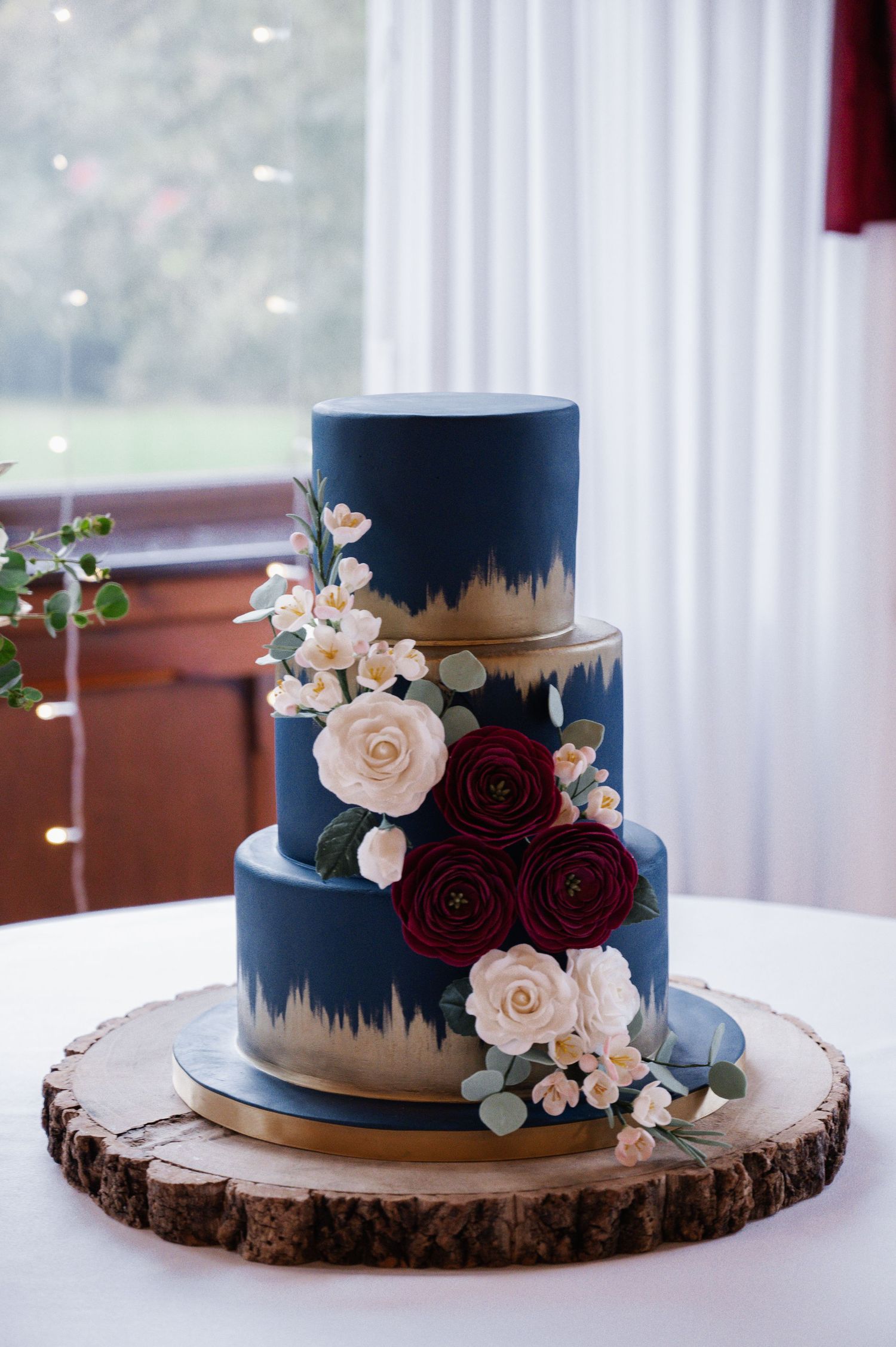 Elegant three-tier navy blue and gold wedding cake decorated with red and white sugar flowers.