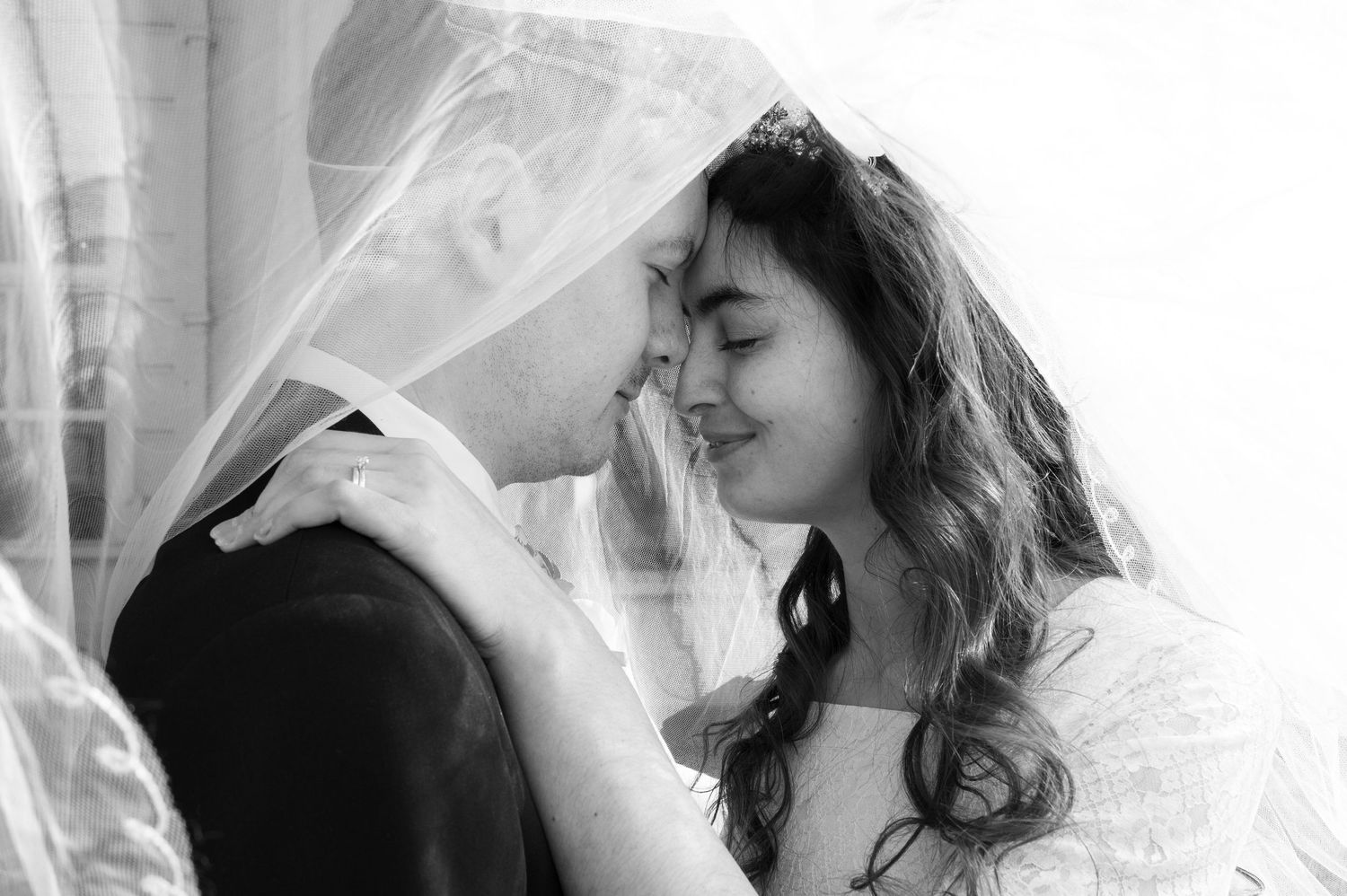 Intimate black and white photo of bride and groom sharing a tender moment under a flowing wedding veil.