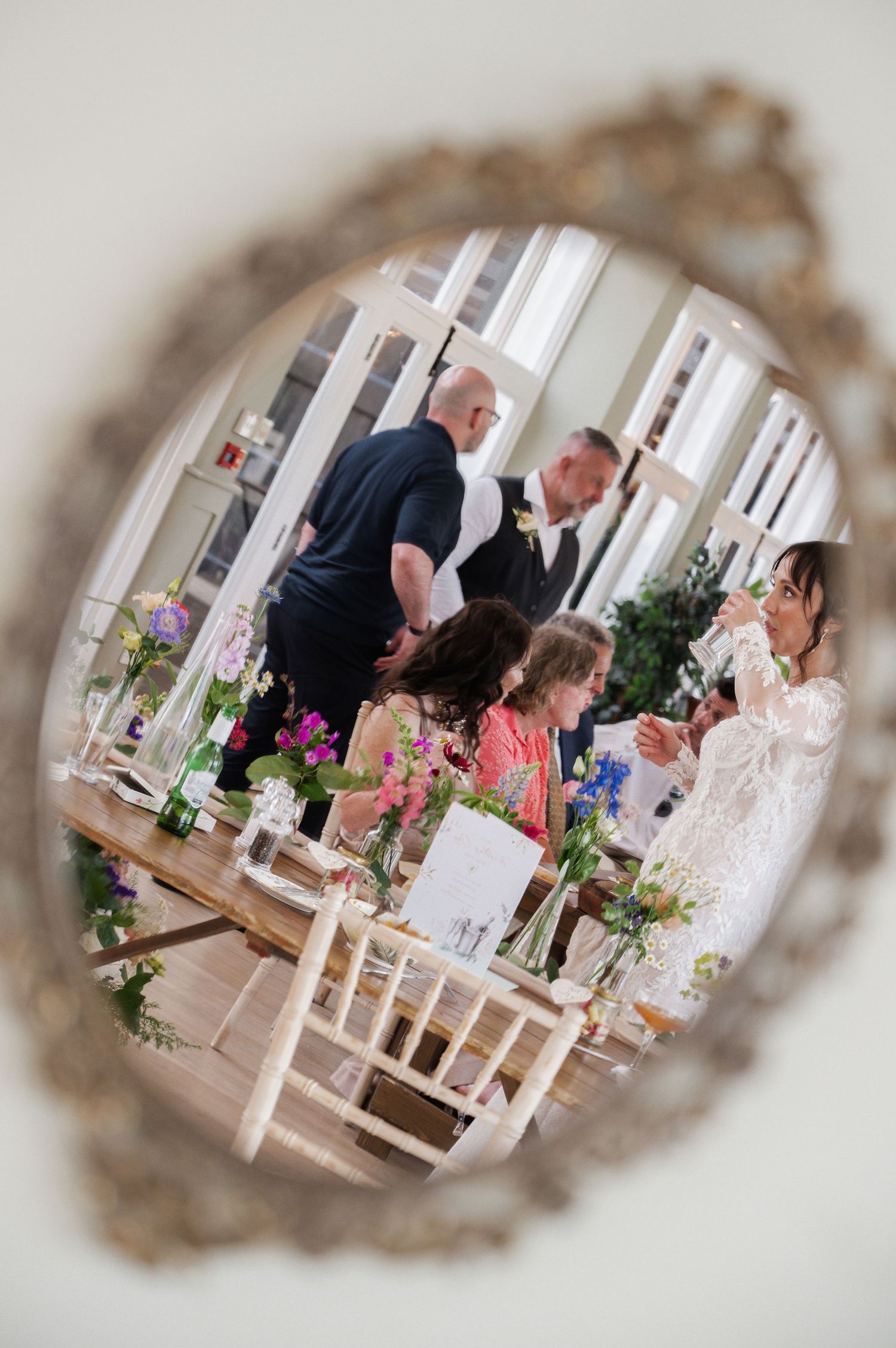 Wedding reception reflected in ornate mirror showing guests at flower-adorned tables in bright venue.