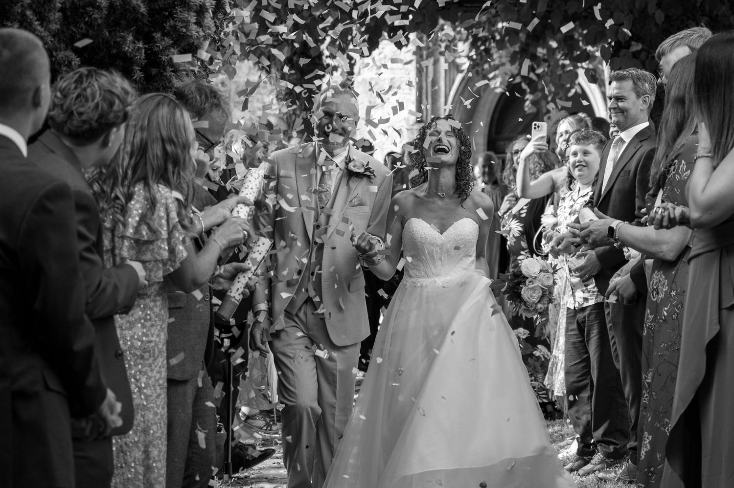 Black and white photo of newlyweds walking through confetti shower outside a church with guests.