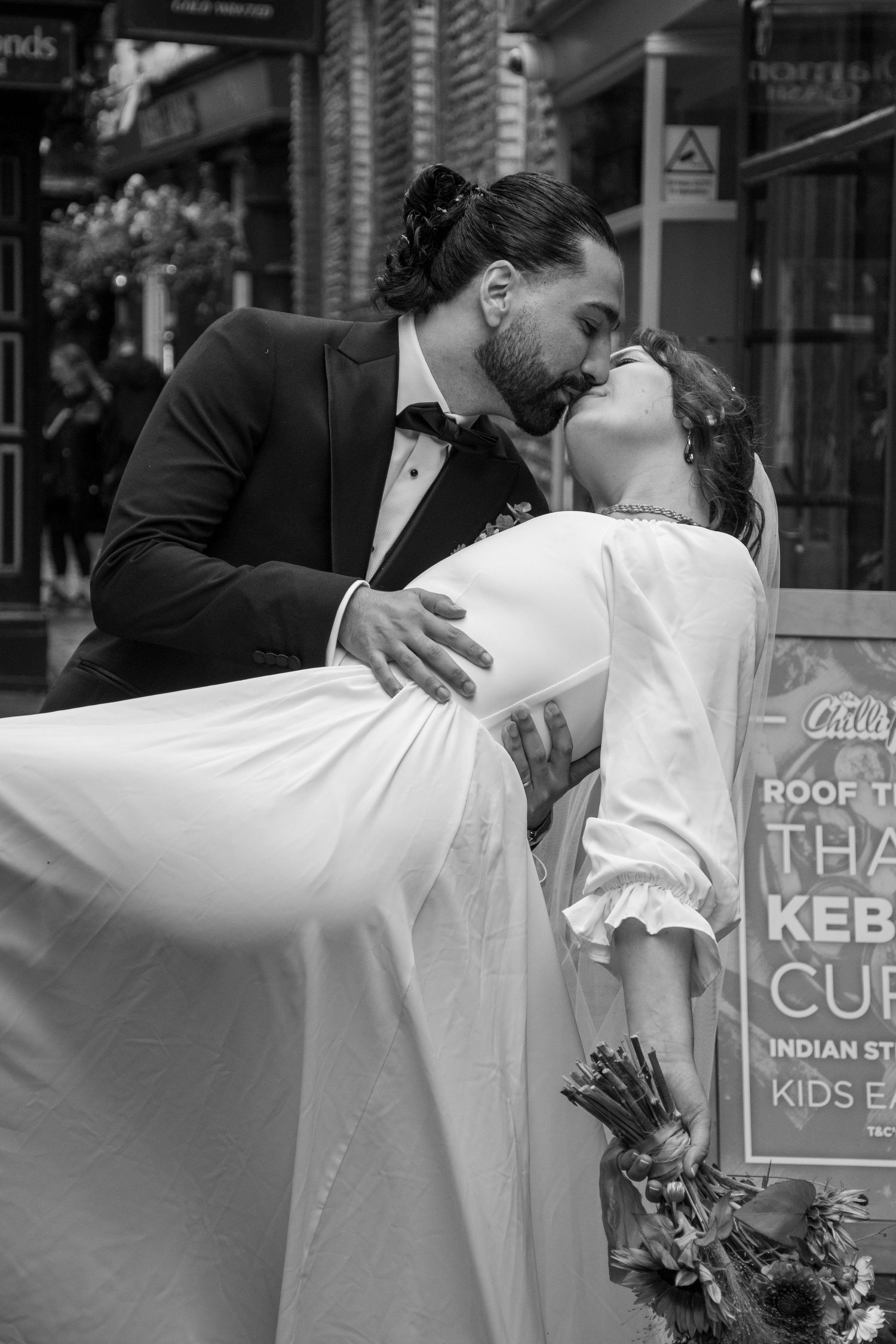 Black and white photo of groom dipping bride for a passionate kiss on a busy urban street after their wedding.