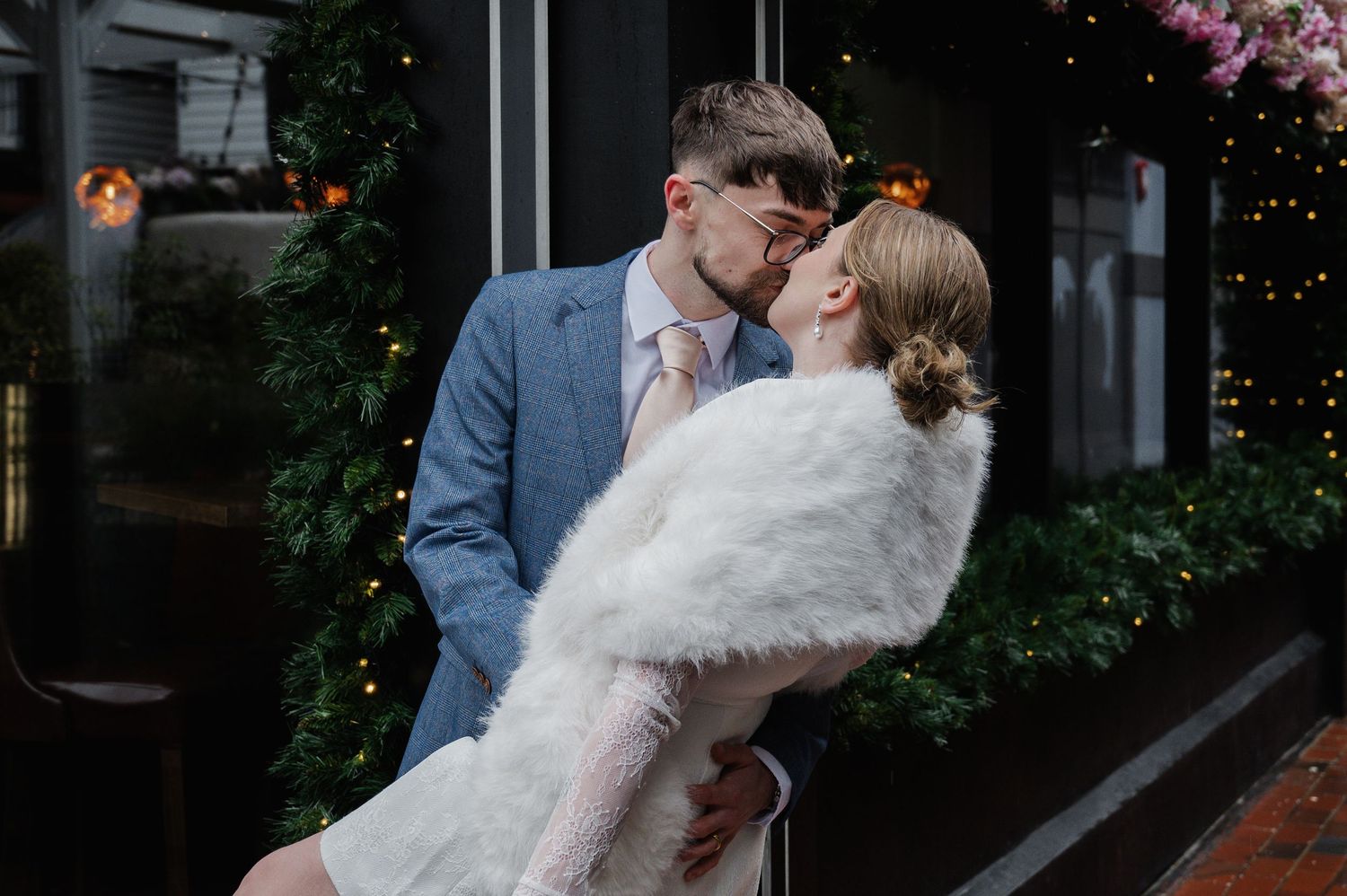 Stylish couple kissing outside a venue decorated with fairy lights and lush greenery, bride wearing a white fur wrap.