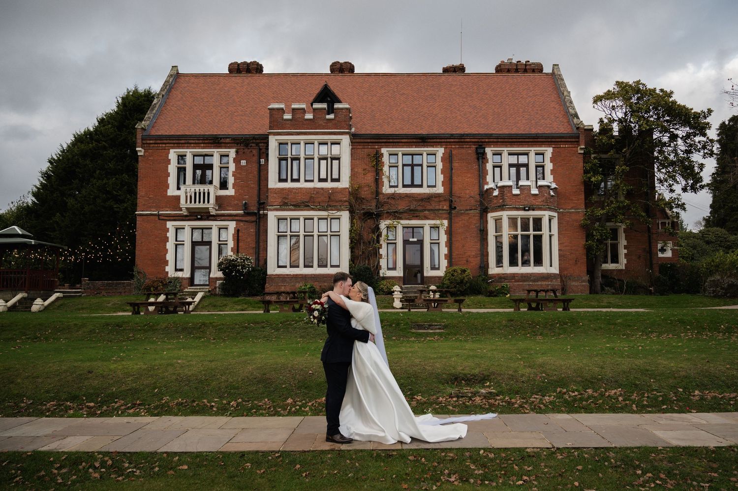 Wedding couple kissing on lawn before grand red brick manor house. Highley Manor