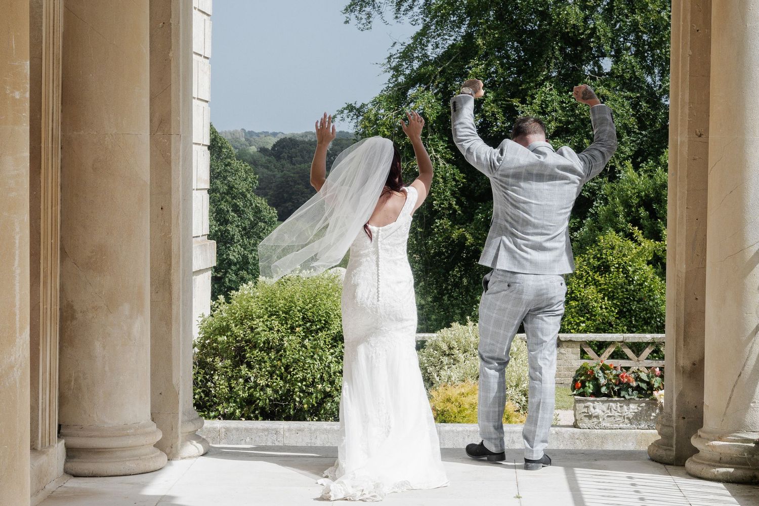 A bride and groom celebrate joyfully between columns with a scenic garden view behind them.