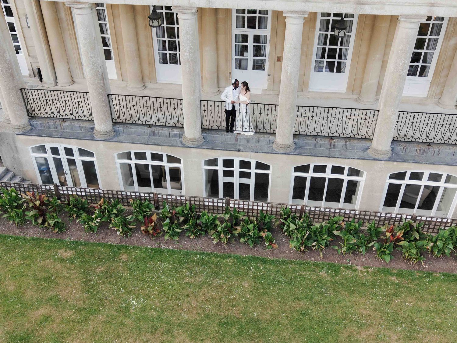 Aerial view of a couple standing on a grand neoclassical building balcony with tall columns and large windows. Buxted Park Hotel