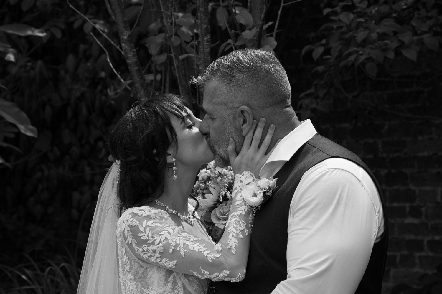 Black and white photo of bride and groom sharing a tender kiss outdoors in lace wedding dress and vest.