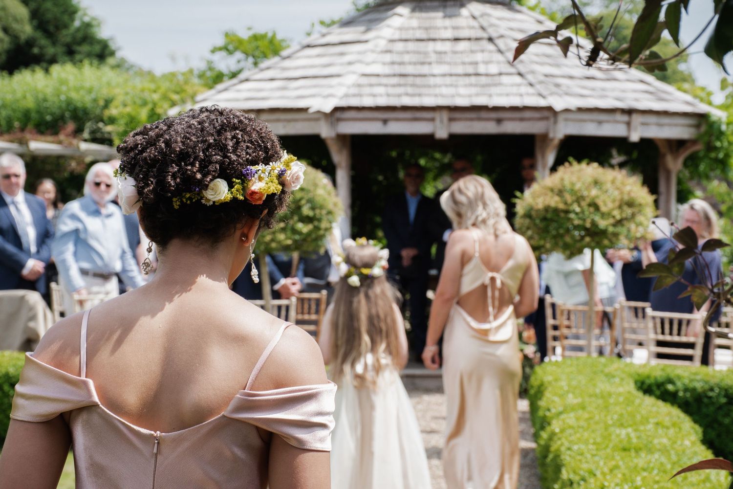 Bridesmaid with floral crown watches bride walk down outdoor garden wedding ceremony aisle.
