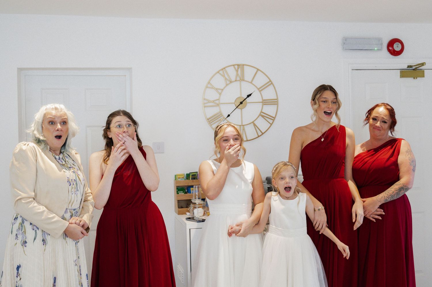 Bridesmaids in red dresses and flower girl reacting with surprise and joy at wedding preparations.
