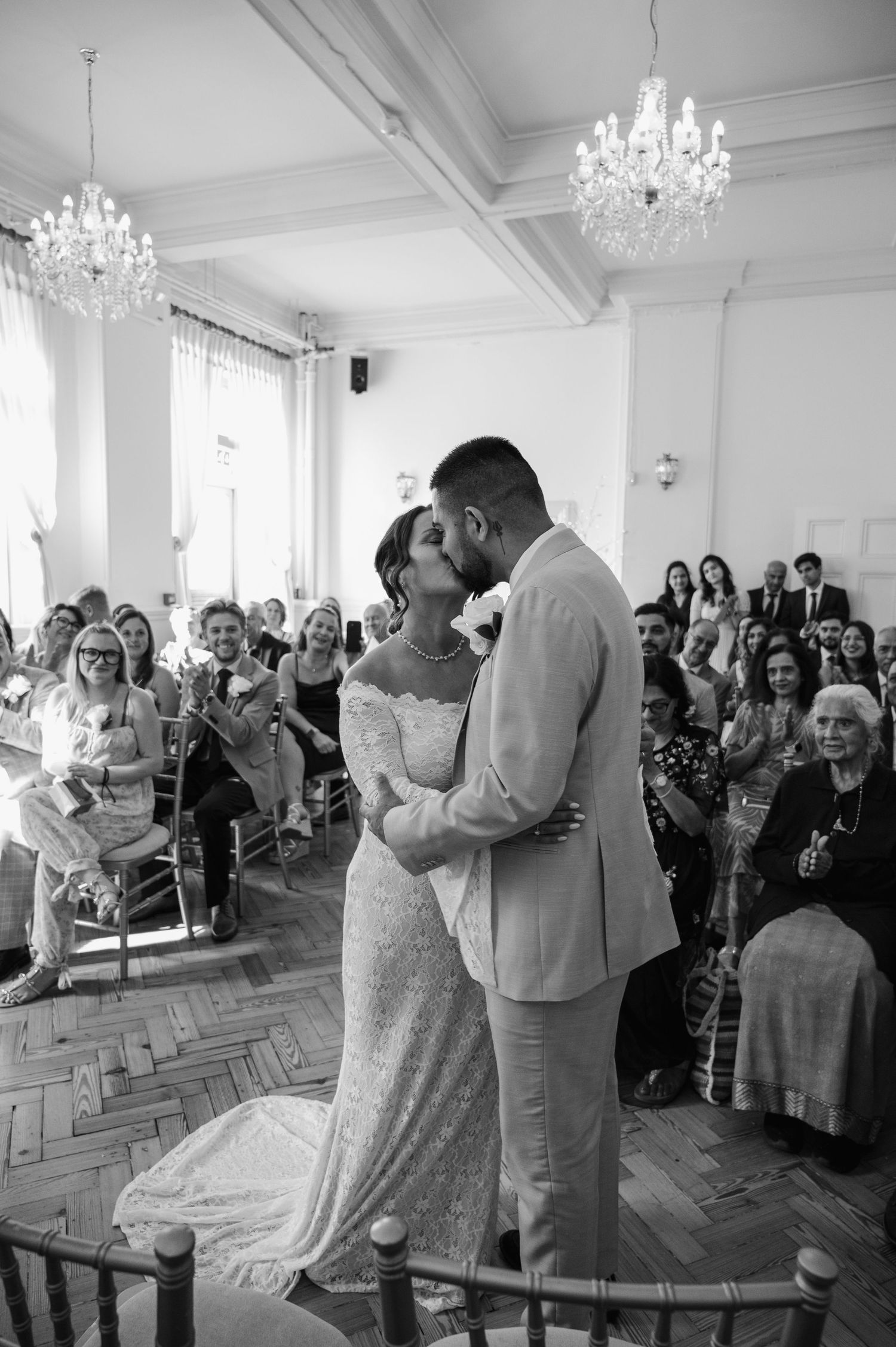 Newlyweds sharing first kiss at civil ceremony under crystal chandeliers with guests applauding.