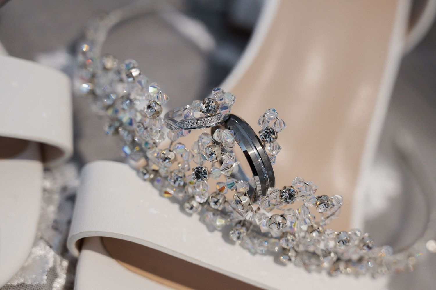 Close-up of silver wedding rings resting on crystal embellished white bridal heels and tiara.