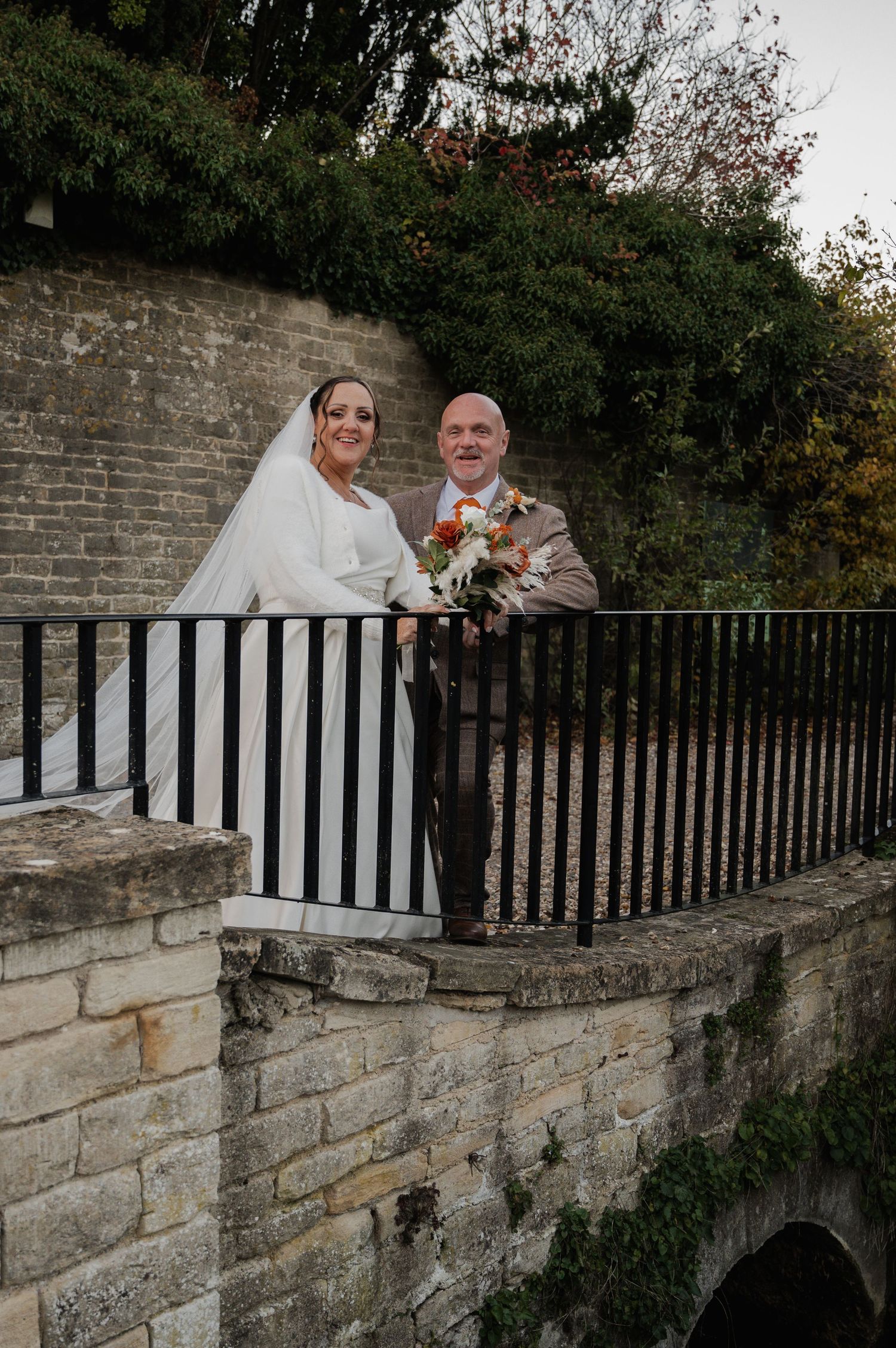 Smiling bride and groom posing together on a stone bridge balcony with autumn foliage at their wedding venue.