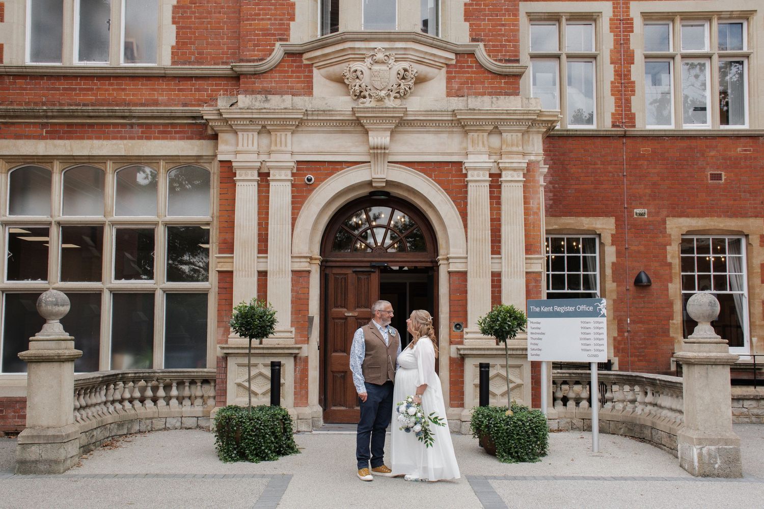 Bride and groom posing before grand red brick building with ornate entrance arch.