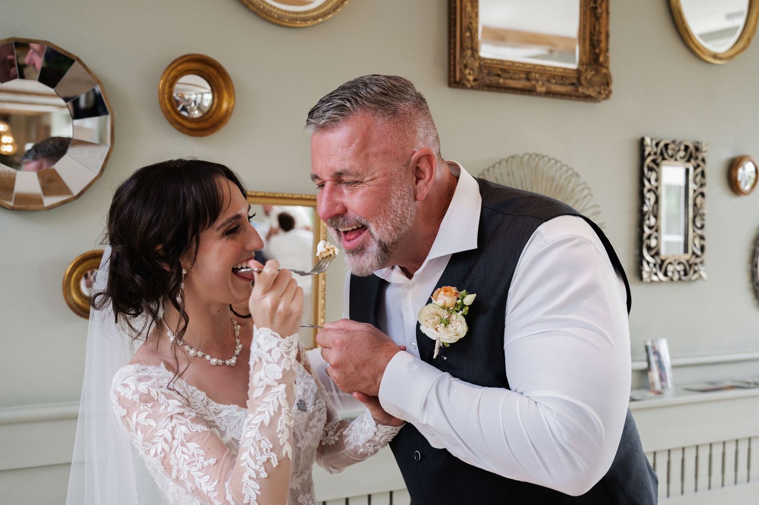 Bride and groom laughing together while tasting wedding cake at their reception indoors.