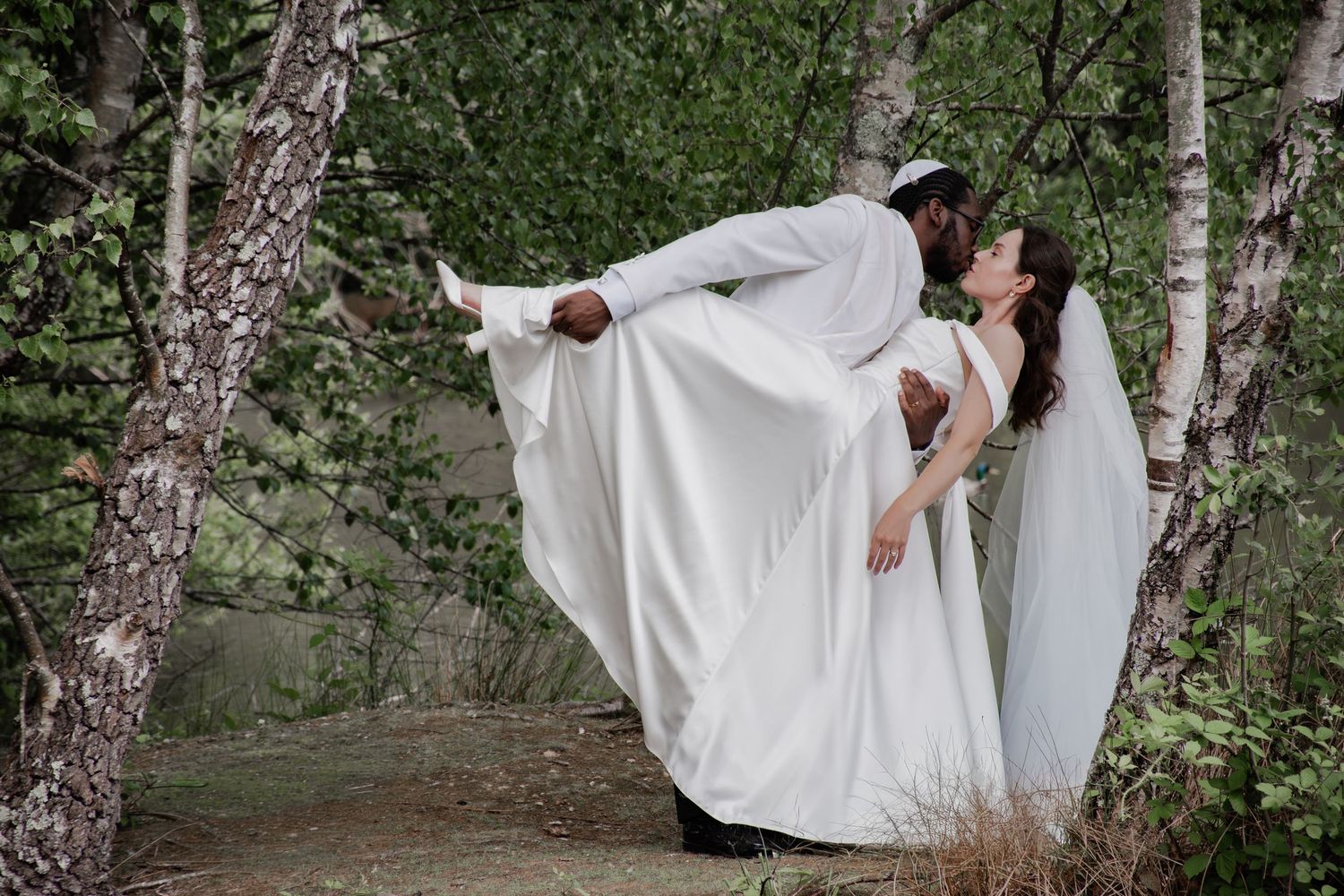 Groom lifting bride in white wedding dress outdoors among birch trees in a romantic forest setting.