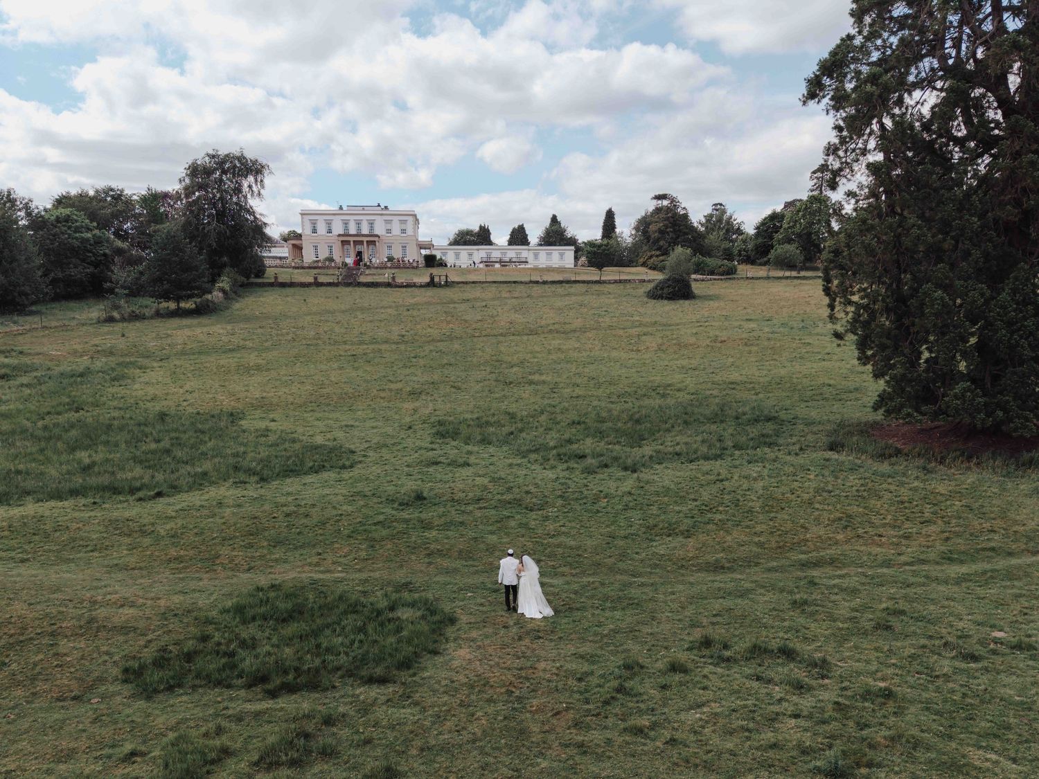 Aerial view of a bride and groom walking across a vast green lawn with a stately manor in the background. Buxted Park Hotel