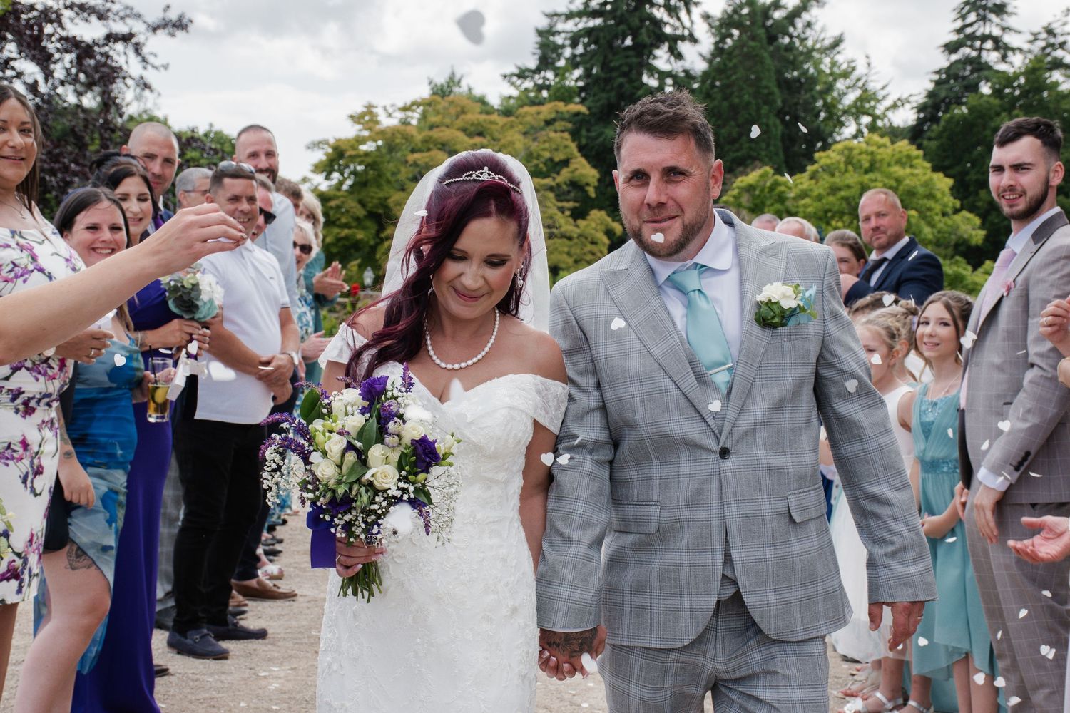 A newlywed couple walks through a confetti shower surrounded by smiling wedding guests.
