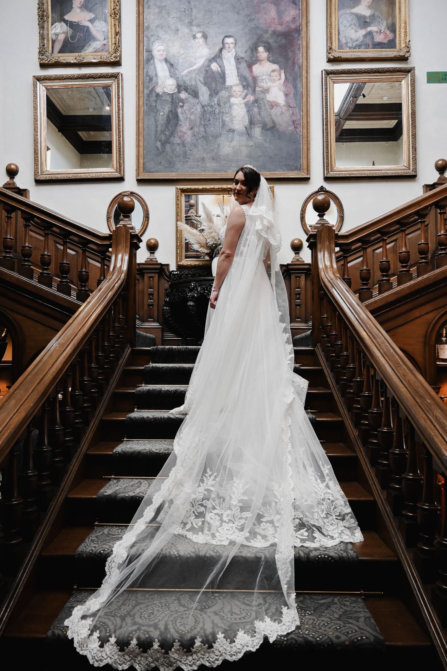 Bride in a lace wedding gown with a long veil standing on a grand wooden staircase.