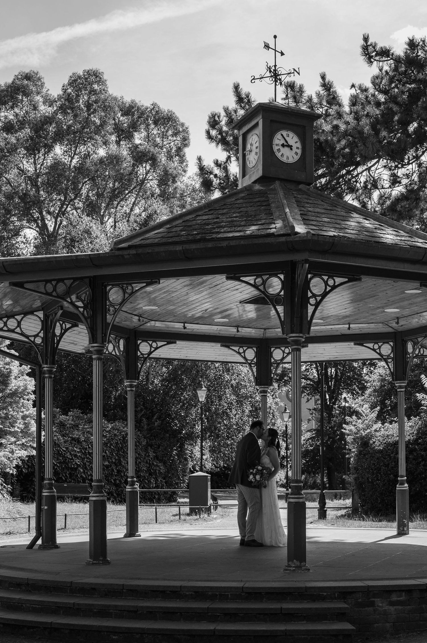 A wedding couple shares a romantic kiss under an ornate Victorian bandstand gazebo with a clock tower in a park.