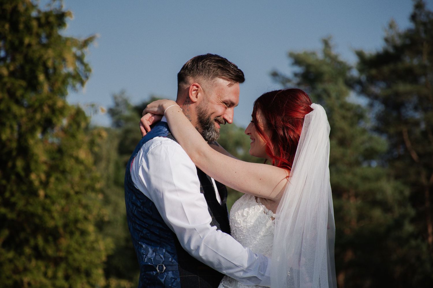 Tattooed groom in blue vest embraces red-haired bride in veil outdoors under blue sky.