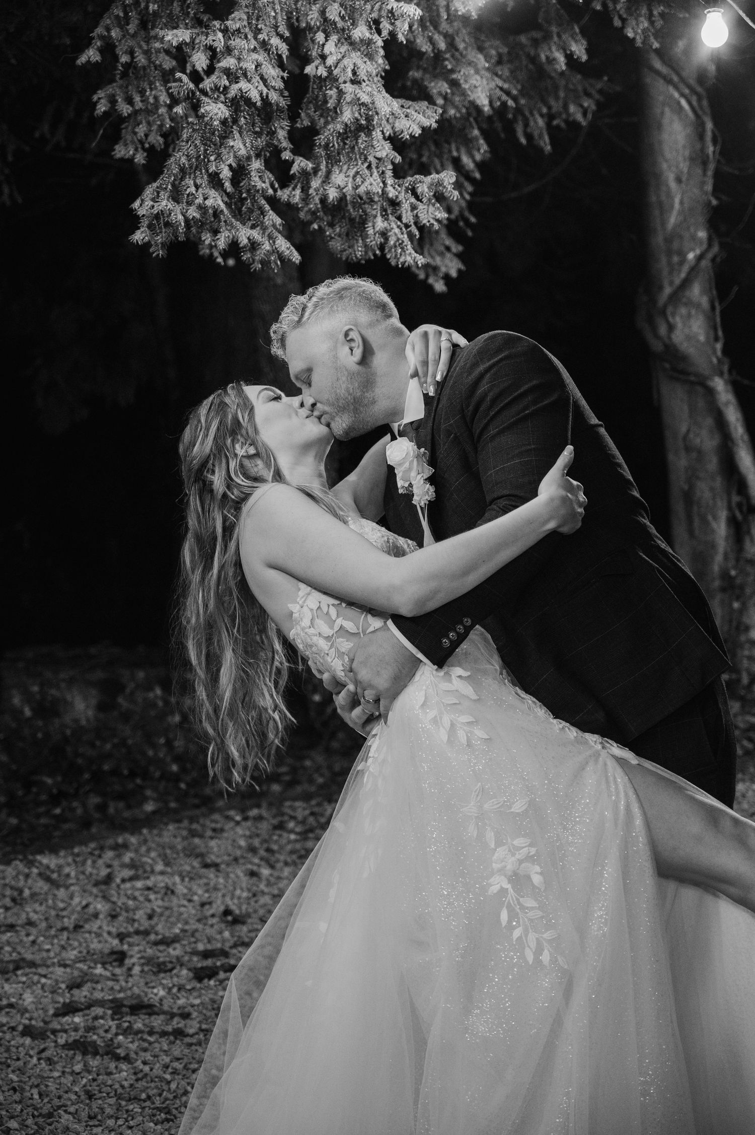 Black and white photo of bride and groom sharing a romantic dip kiss outdoors at their evening wedding celebration.