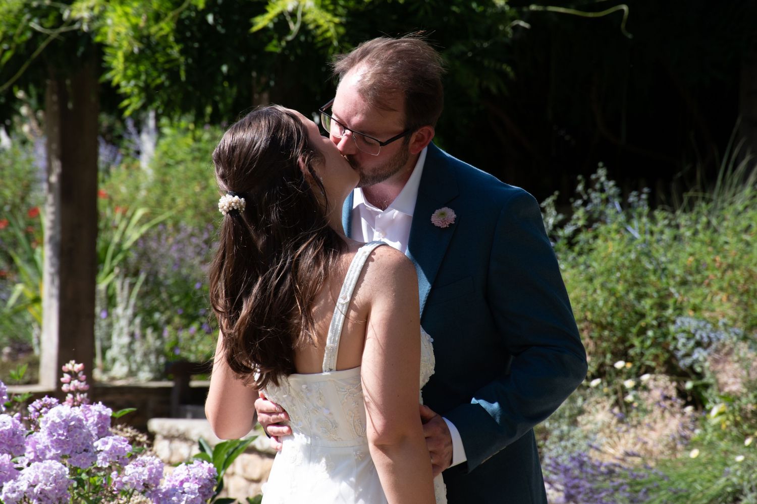 A bride and groom share a tender kiss surrounded by blooming purple flowers in a beautiful garden setting.