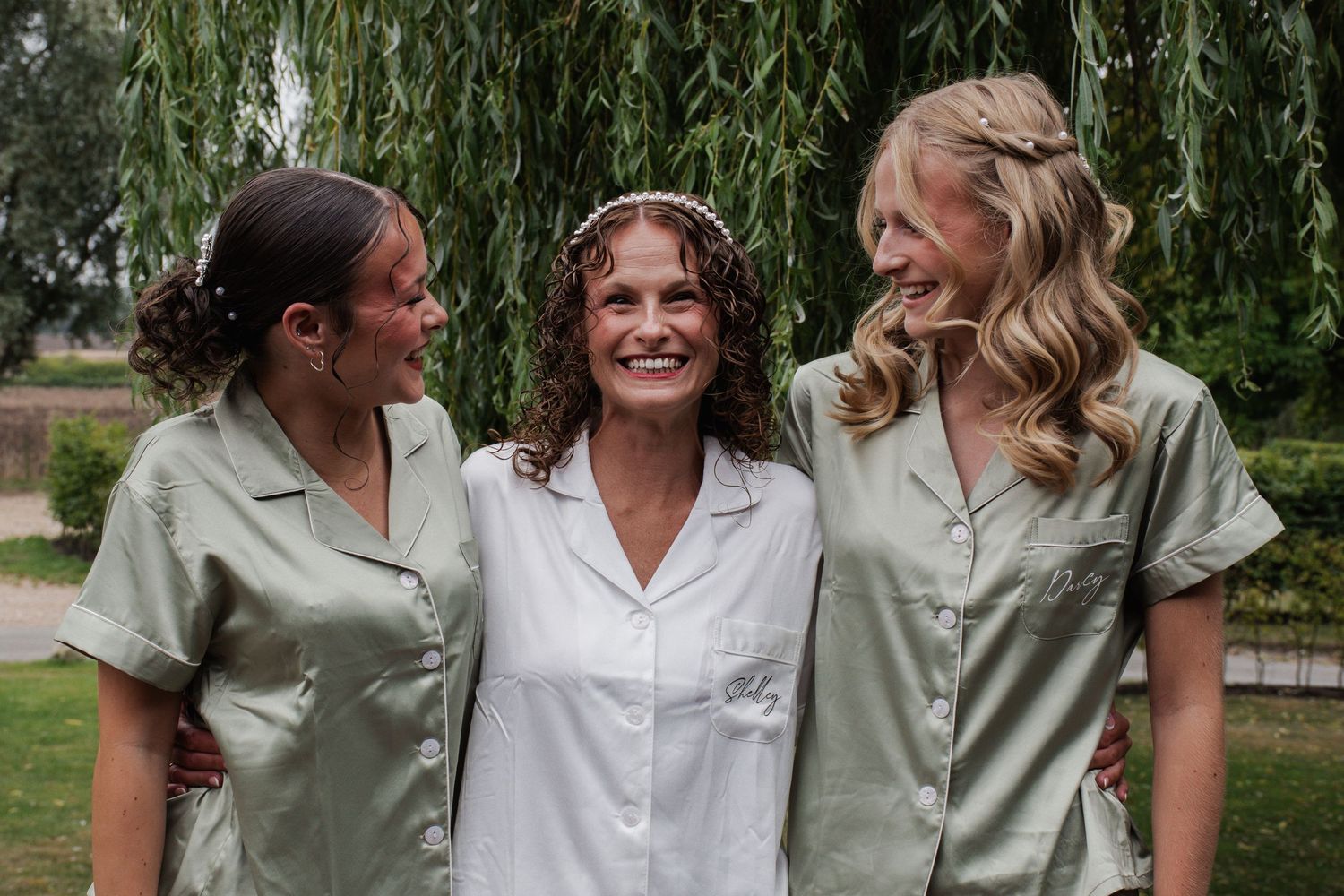 Bride in white robe laughs with two bridesmaids in sage green robes outdoors before wedding.