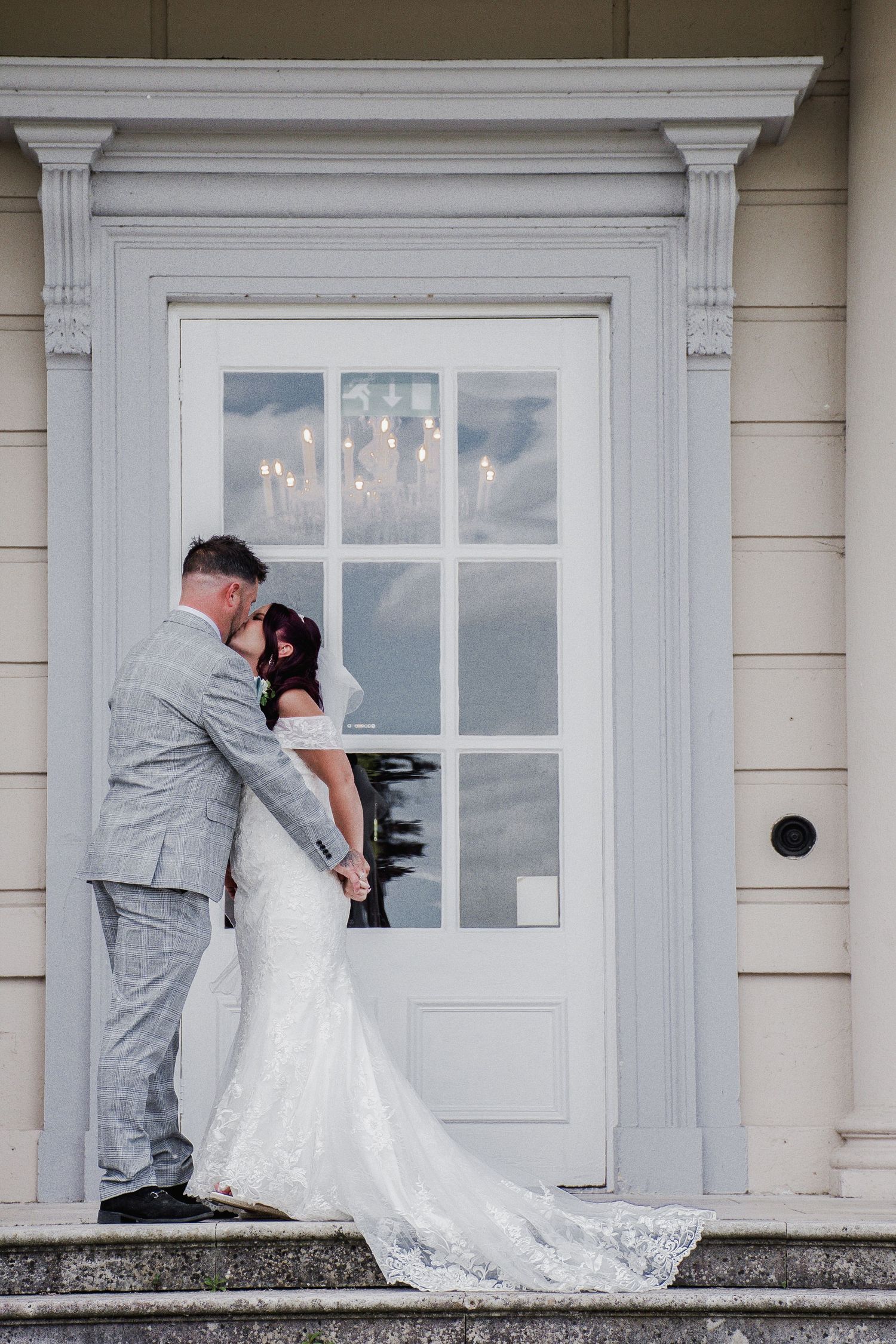 Bride and groom share an intimate kiss on steps before a grand white door with chandelier reflection in the glass.